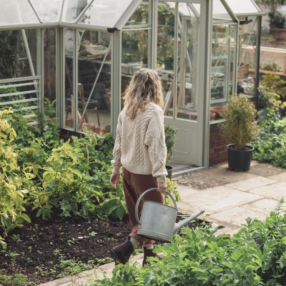 Woman caring a watering can in a garden with a greenhouse in the background