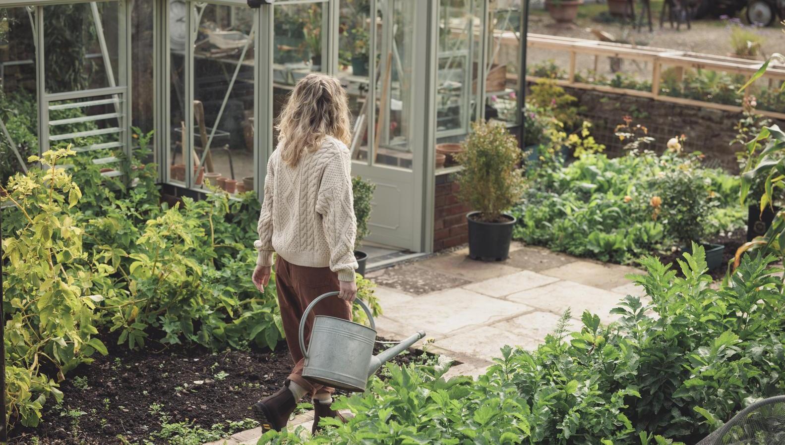 Woman caring a watering can in a garden with a greenhouse in the background